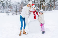 美丽快乐的母亲和女儿微笑着在相机, 而站在雪人在冬季公园