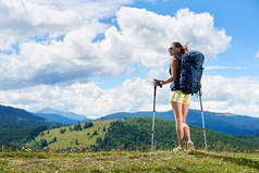 运动员微笑的女子旅游徒步登山小径, 走在草地上, 穿着背包和太阳镜, 使用徒步棍, 享受夏日阳光明媚的一天在山上。旅游理念