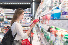 时尚迷人的女人站在冰箱与乳制品, 持有酸奶在他的手中, 并阅读标签。一个有吸引力的女孩在超级市场买酸奶.