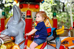 可爱的幼儿女孩骑在游乐园的环形旋转木马动物。快乐健康的孩子在阳光明媚的日子在户外玩得开心。家庭周末或假期
