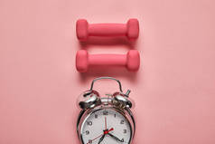 平面躺在银色闹钟和粉红色背景的粉红色哑铃