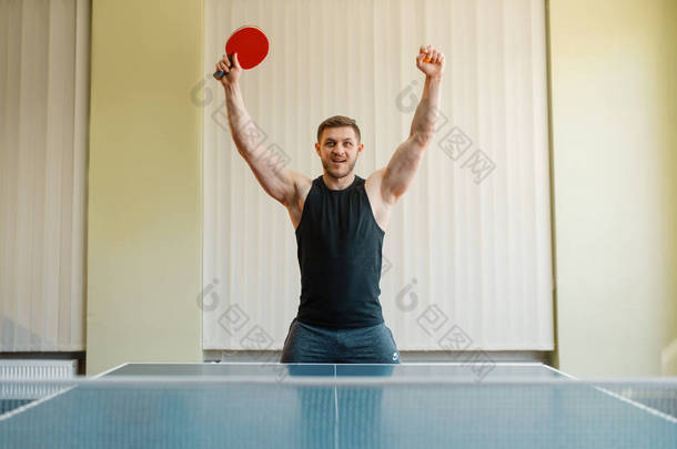 带着乒乓球拍的快乐男人举起双手，在室内比赛。男运动员穿运动服，在乒乓球俱乐部训练