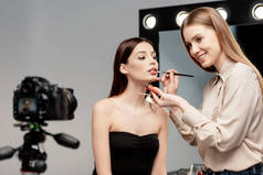 在灰色数码相机旁的模特唇上涂唇彩的快乐化妆画家的选择焦点