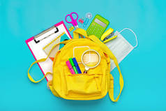回到学校，教育理念黄色背包，配有学习用品、防护性医疗面罩、计算器、剪子等蓝色背景材料。顶视图复制空间平铺
