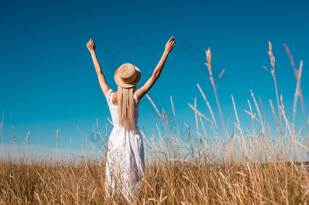 穿着白衣、头戴草帽的时髦女人站在青草丛生的草地上，<strong>高举</strong>双手在蓝天衬托下的背影