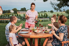 夏天外出野餐时,一家人在自家花园的烤架边吃饭.围坐在餐桌旁，端着食物和碗碟的人