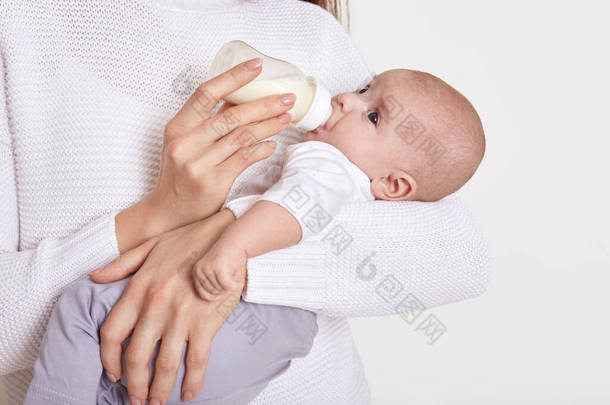 妈妈用奶瓶喂她的孩子，她的孩子被白色的背景隔离了，小宝宝在妈妈的手里，她吃着牛奶，孩子穿着白衬衫和灰色的裤子。童年和母亲.