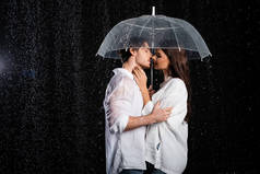 年轻迷人的男女朋友站在雨中，带着雨伞，吻着黑色的背景
