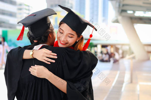 两个戴着方顶学术帽的快乐的毕业女生互相拥抱，人们庆祝教育的成功，并在大学毕业典礼上和朋友一起祝贺他们毕业.