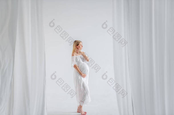 一个年轻的孕妇在期待着一个穿着白衣的孩子，抱着她的肚子。时尚女孩在一个简约的，孤立的内部削减空气的面料。容易、快乐的孕产、代孕.