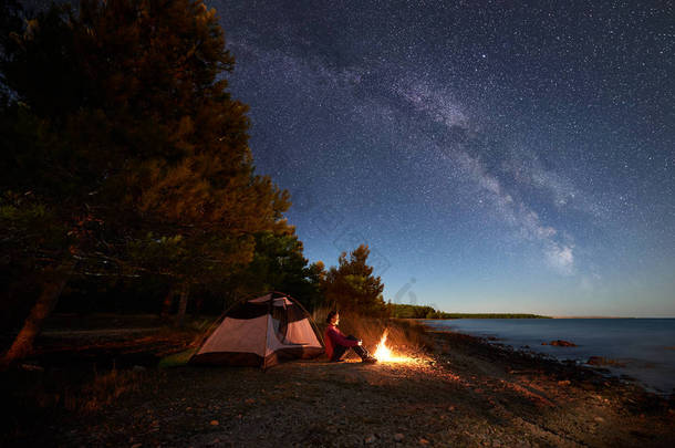 在海边过夜露营。女徒步旅行者坐在帐篷前的篝火旁, 在明亮的星空和银河下, 享受着美丽的蓝色<strong>水景</strong>色。旅游与积极的生活方式理念