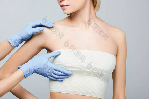 裁剪视图整形外科医生在乳胶手套检查女性乳房绷带, 隔离在灰色