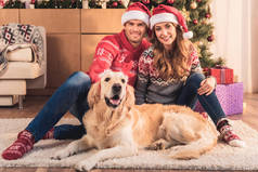 美丽的情侣在圣诞老人的帽子坐在圣诞树与猎犬狗在鹿角