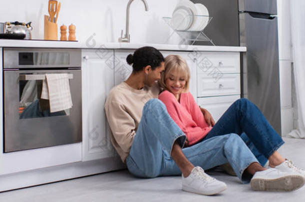 positive multiethnic couple hugging and sitting on floor in modern kitchen, <strong>banner</strong>