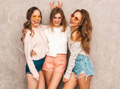 三个年轻美丽的微笑的嬉皮士女孩在时尚的夏季运动服装。性感无忧无虑的女人摆在灰色的墙附近。穿着圆形太阳镜的模特玩得很开心。用手指在她头上做角
