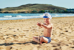 快乐的孩子坐在沙滩上玩沙倒在他的手。漂亮的小男孩微笑放松在假期，温暖的夏日，户外。背景海岸，海洋，自然。度假，度假，生活方式.