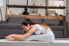 在客厅里, 在家里的健身垫上练习坐着向前折叠姿势的妇女