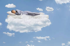 穿着睡衣睡在床垫上，飘浮在云彩和蓝天之上的人