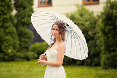 美丽的新娘在白色礼服与装饰伞反对背景绿色自然。在一个夏日公园里