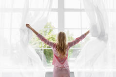 穿着粉色睡衣的金发年轻女子醒来，看着卧室里的大玻璃窗