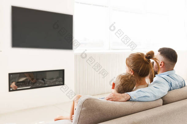 年轻的家庭、父亲、母亲和两个小孩坐在壁炉前的沙发上看<strong>电视</strong>，在家里聚精会神地享受闲暇时光的<strong>背景图</strong>