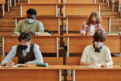 在大学的课堂上，一群戴着防护面具的学生坐在长椅旁，一边做着个人任务