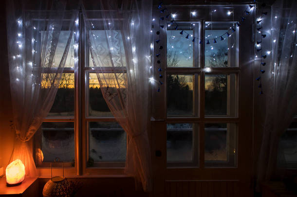 装饰的喜马拉雅山盐灯被白色网布窗帘的古老的白色木制窗户打开，冬季的时候，从木制框架窗户向外眺望着森林的落日。冰封的玻璃.