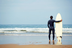 有吸引力的年轻冲浪者抱着冲浪板，而站在海滩上看着海洋，找到一个完美的位置去冲浪海浪，在专业的冲浪者，以冲浪板看着海洋，过滤图像