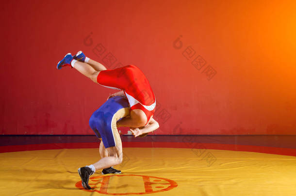两名身穿<strong>红蓝</strong>相间制服的男子摔跤选手在体育馆的一块黄色摔跤地毯上摔跤