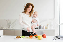 美丽的母亲的肖像抱着她可爱的小宝宝, 而站在厨房和烹饪隔离
