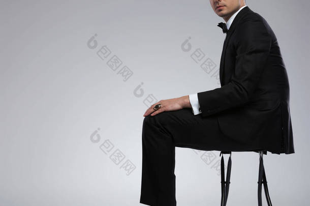 穿着黑色燕尾服的坐着时髦的年轻人的侧面在浅灰色背景, 有一双闪亮的黑色鞋子和一个金戒指在手指上