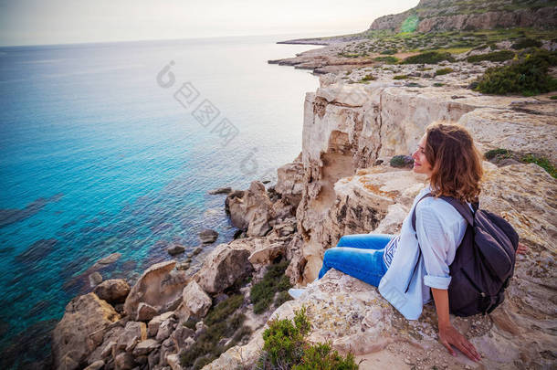 一个时髦的年轻女子旅行者观看海滩上的岩石上美丽的日落, 塞浦路斯, 希腊角, 欧洲夏季旅游的热门目的地。
