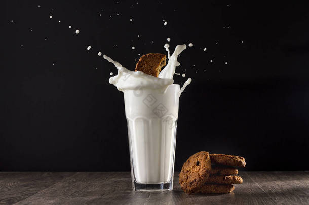 燕麦饼干和玻璃牛奶在一个黑色的背景木桌上。一块饼干落入玻璃杯中.