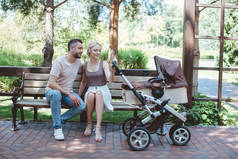 父亲和母亲坐在长椅上在公园的婴儿马车附近