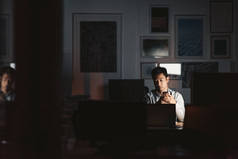 年轻的亚洲商人深夜坐在黑暗的办公室里, 在一台笔记本电脑上工作, 喝着一杯咖啡。