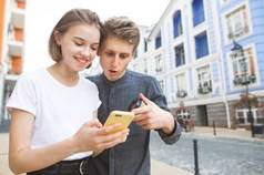 美丽的年轻夫妇使用智能手机在城市的街道上。女孩看着智能手机和微笑, 年轻人显示手指。两个游客正在看智能手机上的路线.