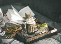 秋季或冬季热巧克力或咖啡与奶油和肉桂在乡村杯在木托盘在床上, 背景深色墙壁, 水平组成。秋暖甜饮