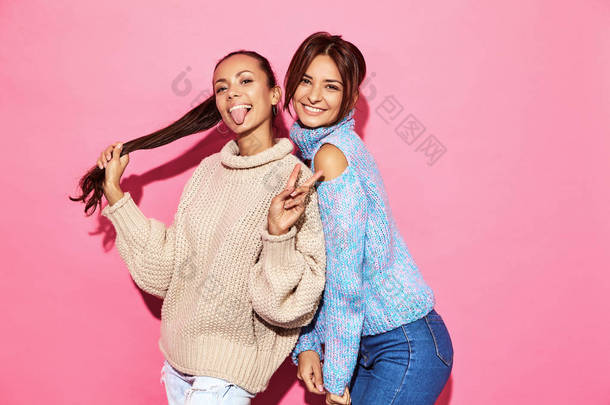两个美丽性感的微笑美丽的女孩看着相机。热妇女站在时尚的白色和蓝色毛衣, 粉红色背景。显示和平标志