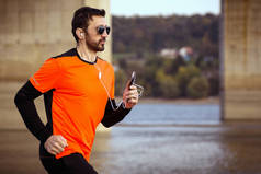 The fit man in orange sportswear is jogging outdoors with earphones and phone, recreation concept.