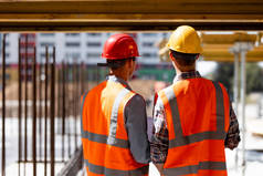 两名身穿橙色工作服、头戴头盔的土木工程师在木结构和钢架附近的建筑工地上讨论施工过程
