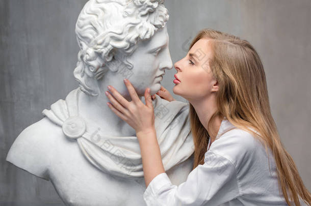 年轻女子亲吻上帝阿波罗半身像雕塑。古希腊太阳神和诗歌石膏副本上的大理石雕像在格兰格混凝土墙体背景上。古代艺术与生活之美