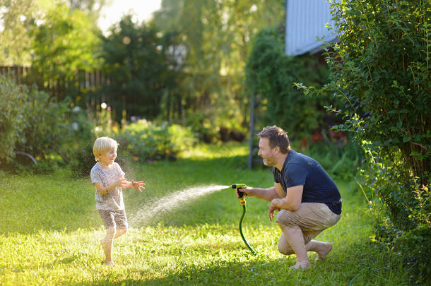 有趣的小男孩与他的父亲玩花园软管在阳光明媚的后院。学龄前儿童在用<strong>水喷雾</strong>玩乐.
