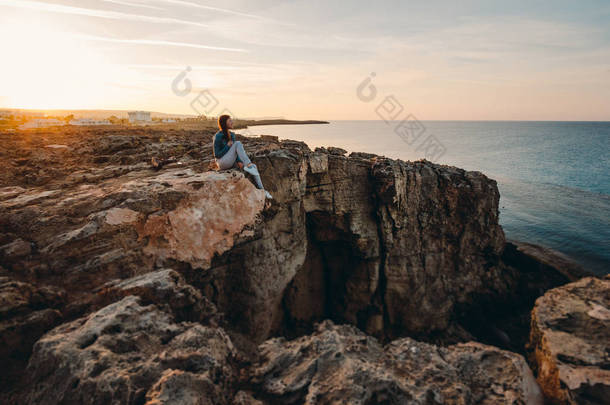 在夏天的群山中，女旅行家坐在悬崖顶上的海边，欣赏着大海和大自然的美景。 希腊角，塞浦路斯，地中海。 日出