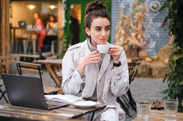 在街上的咖啡店里，穿着风衣的漂亮随意的黑发女孩在笔记本电脑上喝咖啡