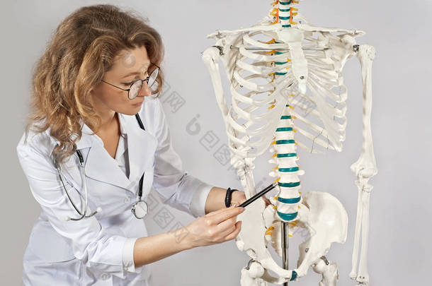 穿着医疗服的女人 医生的骨骼布局。 戴眼镜的女医生隔离在白色背景下. 医生的助手指向骨头. 肌肉骨骼系统疾病的研究.