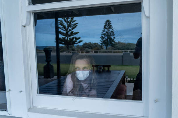 COVID-19封锁。在隔离期间，一个戴着面具的忧郁寂寞小女孩从窗户往外看。可悲的病童在家里孤立无援.Coronavirusu疫情与儿童心理健康.