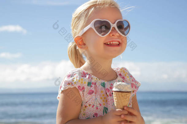 可爱的幼儿女孩吃冰淇淋。孩子们戴着太阳镜，手里拿着一个有美丽蓝色大海和蓝天的冰淇淋的肖像。在海滩度过了一个愉快的暑假.