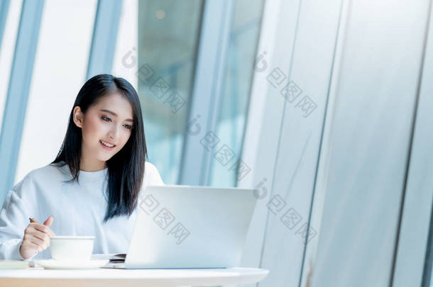 创业者企业主有魅力的亚洲女性商人女人与智能手机和笔记本电脑的交流，白色连衣裙开开开心心，面带微笑，办公室背景模糊不清