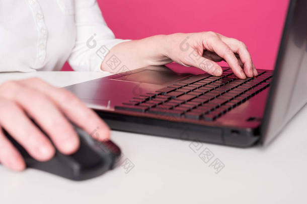 把一个老年妇女的手放在鼠标和笔记本电脑键盘上.她玩电脑游戏或上网搜寻，在社交媒体网站聊天。白色桌子和粉色背景