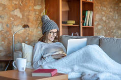 笑着漂亮的年轻女子坐在沙发上和笔记本电脑一起工作。她戴着一顶羊毛帽子和一副眼镜.横向图像